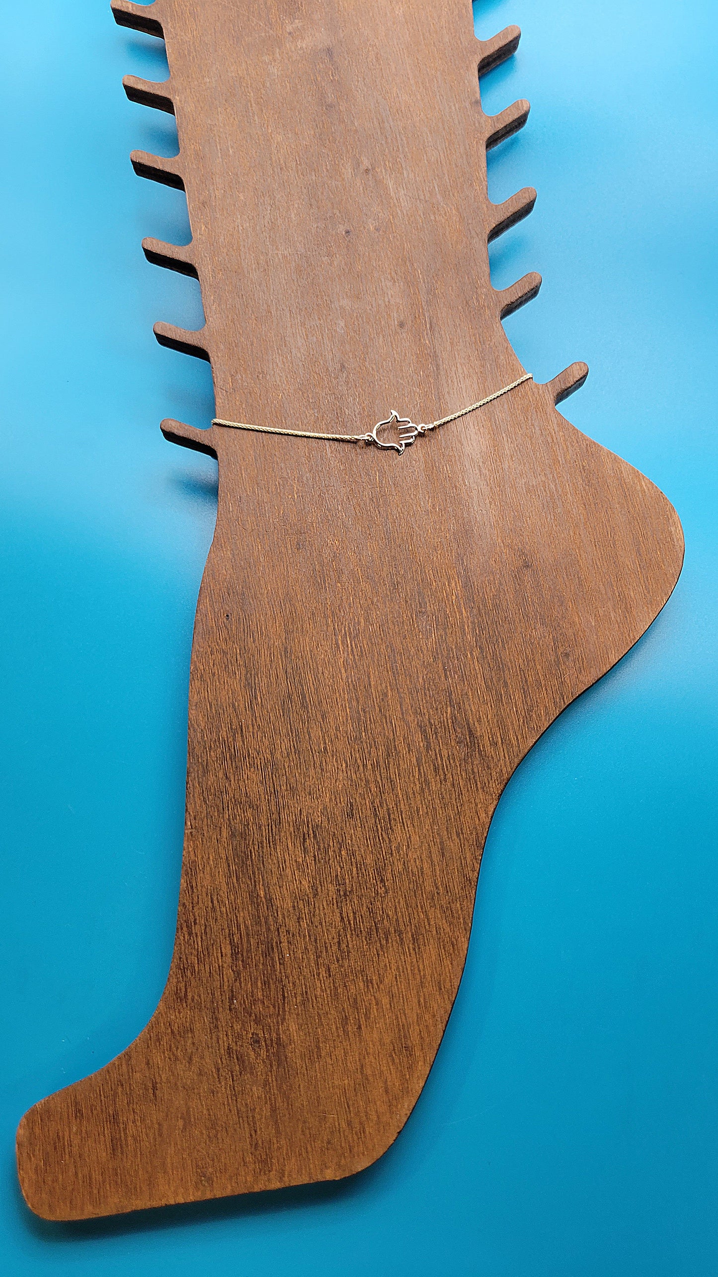 14k gold Hamsa hand Anklet on a wooden foot display while on a blue color background at distance.