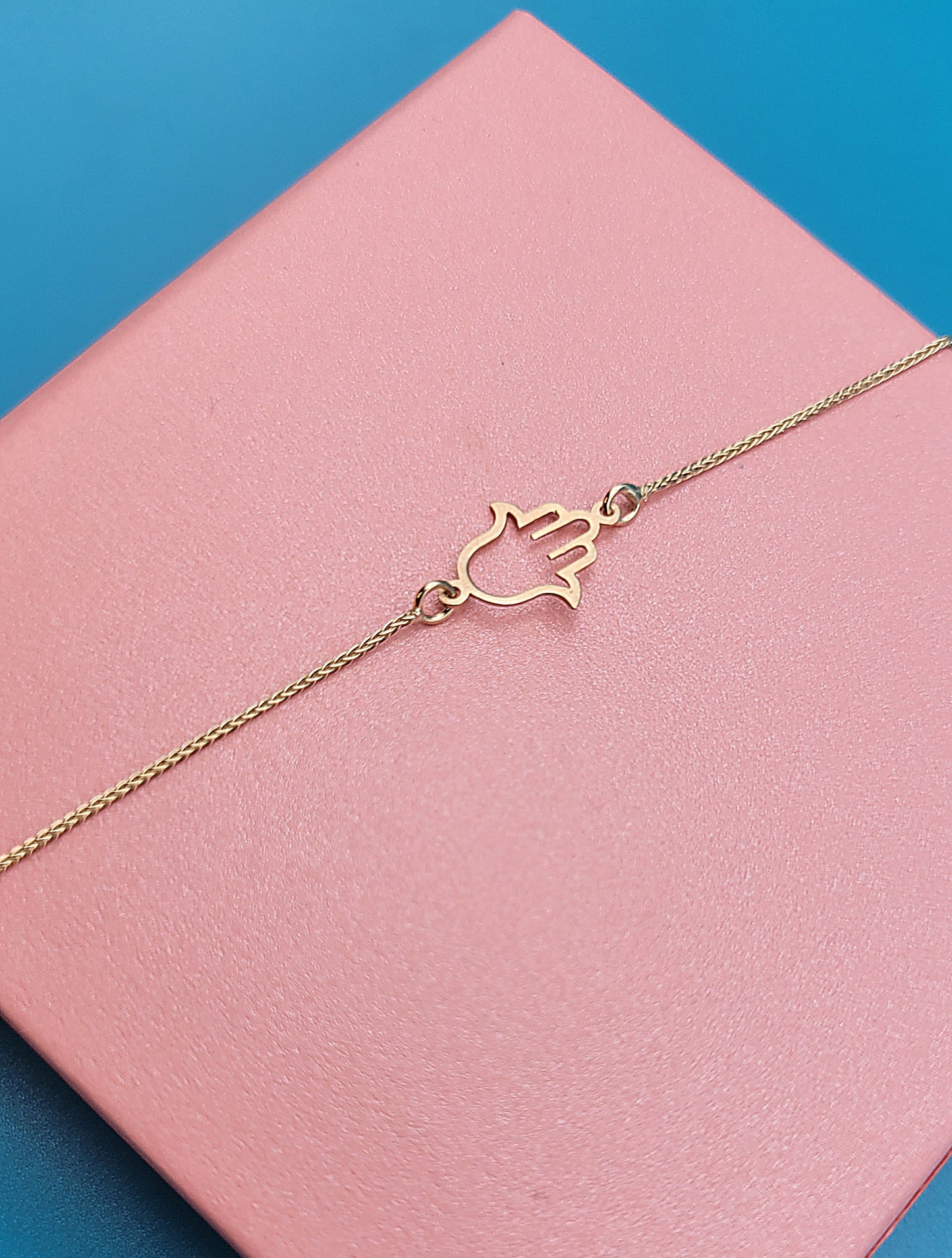 Our stunning 14k yellow gold Hamsa Hand Bracelet displayed angled on top of a pink Jewely box on a blue color background.