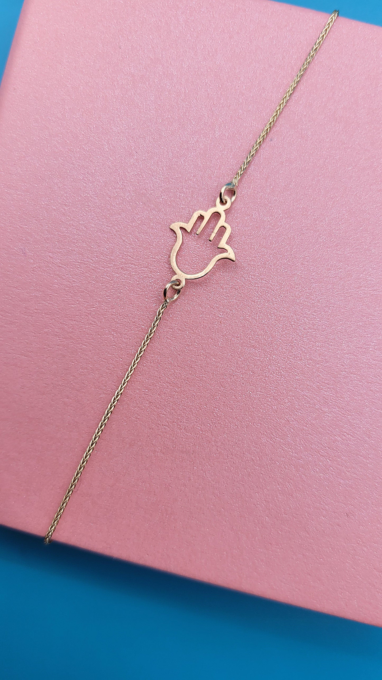 14k yellow gold Hamsa hand bracelet on a pink jewelry box zoomed, blue background.
