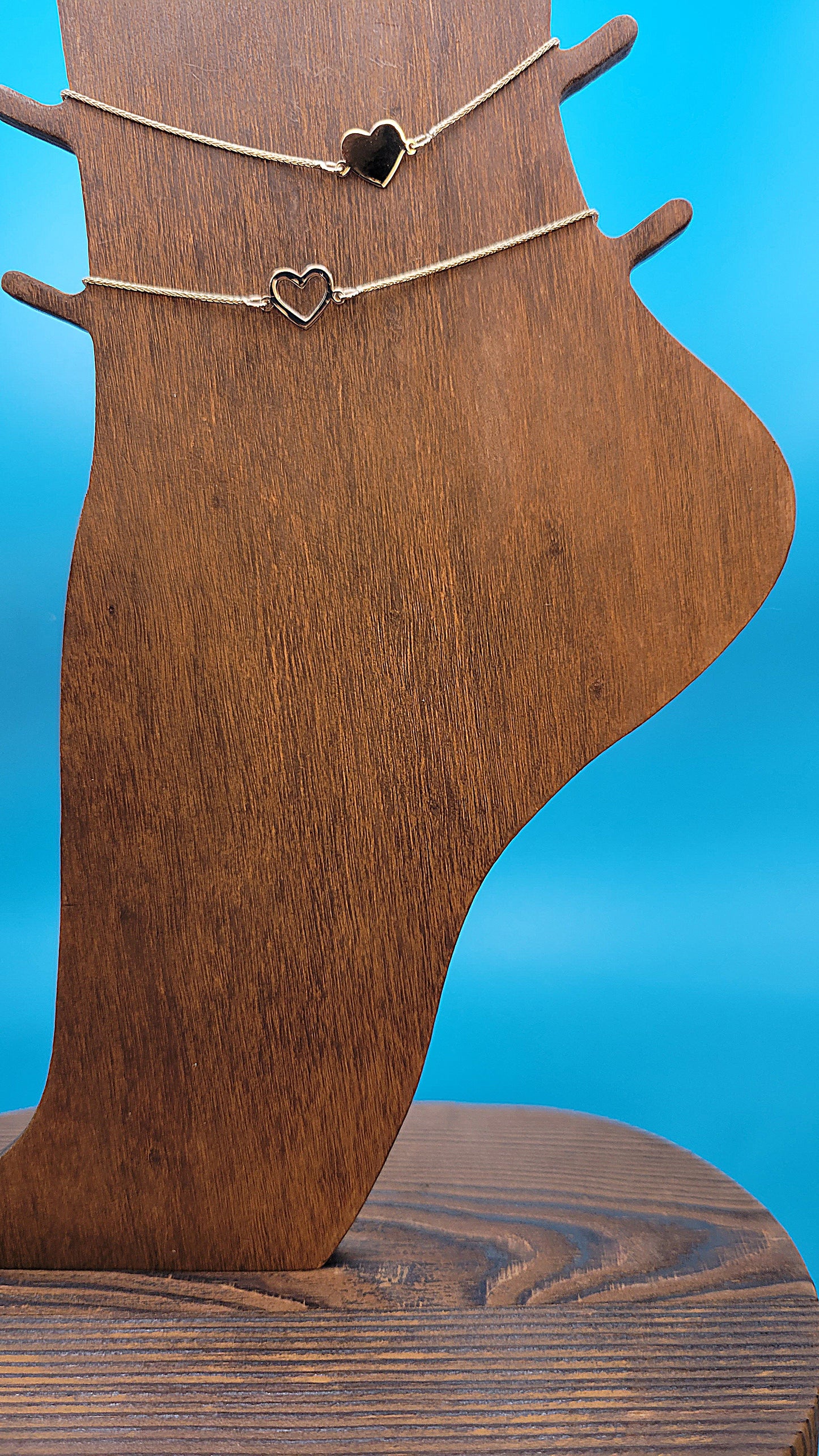 14k gold Hollow and Solid Heart Anklets on wooden foot display on blue color background zoomed in.