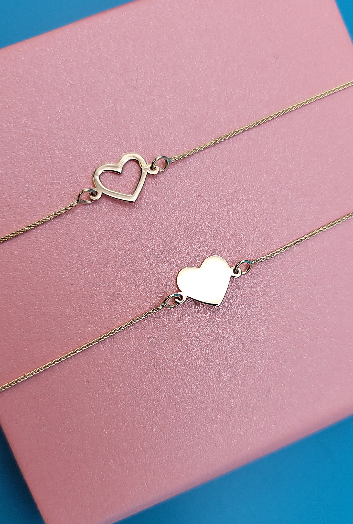 14k yellow gold hollow and solid heart bracelets on a pink jewelry box zoomed, blue background.
